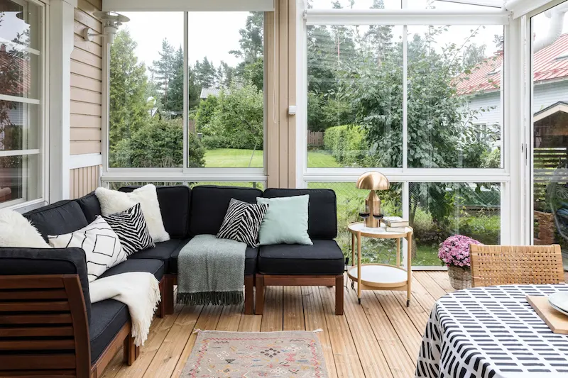 Cozy sunroom with black sectional sofa, decorative pillows, and enclosed balcony-style windows overlooking a garden.