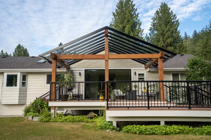 Elevated deck with glass railing and pergola-style roof systems, overlooking a landscaped backyard garden.