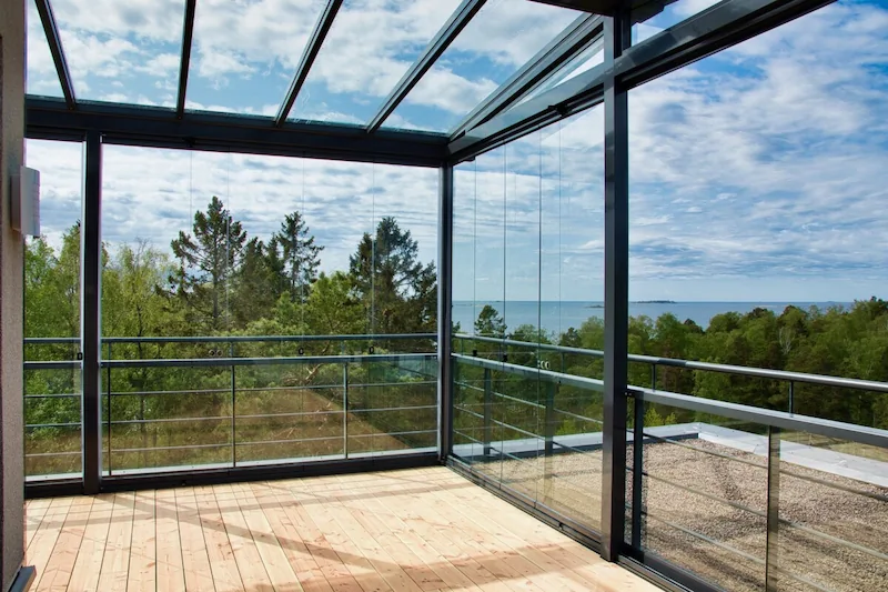 Balcony enclosure with frameless folding glass walls offering unobstructed views of the surrounding forest and ocean.