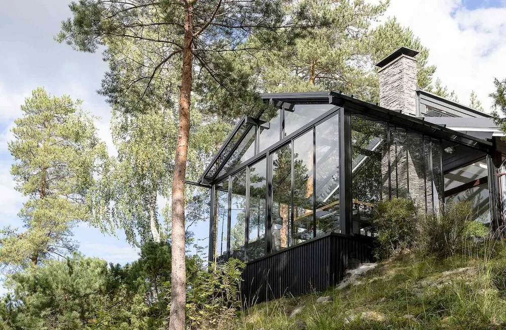 Contemporary hillside home featuring a glass sunroom and balcony enclosure surrounded by forest.