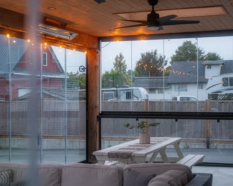 Modern covered patio with frameless glass walls and integrated railings, featuring string lights, a ceiling fan, and a picnic-style dining table.