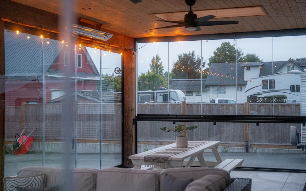 Modern covered patio with frameless glass walls and integrated railings, featuring string lights, a ceiling fan, and a picnic-style dining table.