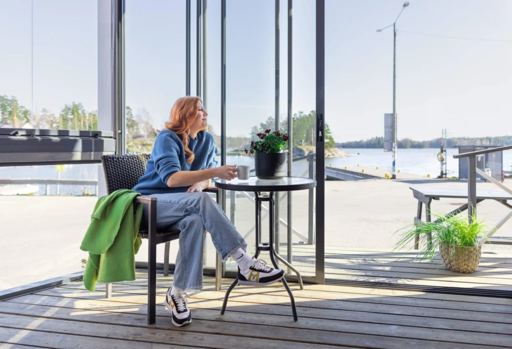 Woman enjoying coffee inside a modern glass sunroom enclosure with a waterfront view.
