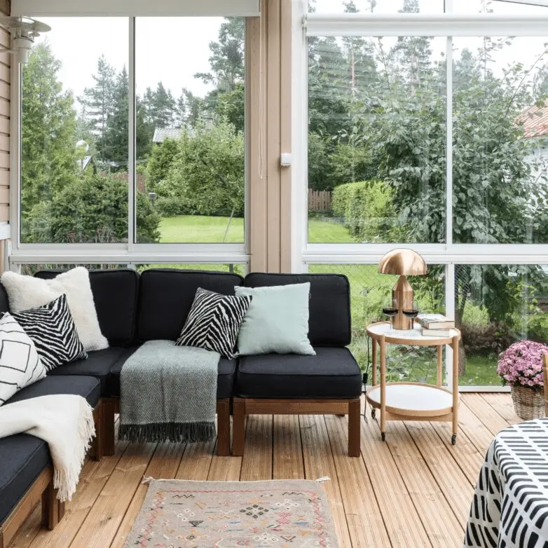 Cozy enclosed sunroom with black sectional sofa, patterned pillows, and large glass windows overlooking a backyard.