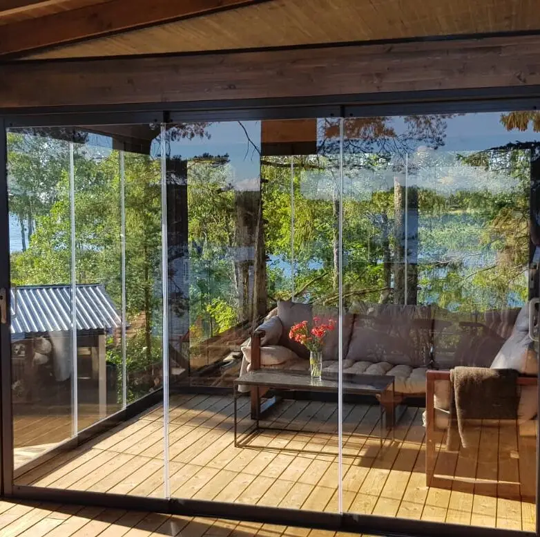 Patio enclosure with glass walls surrounded by forest views, with wooden decking, sofa seating, and a coffee table decorated with flowers.