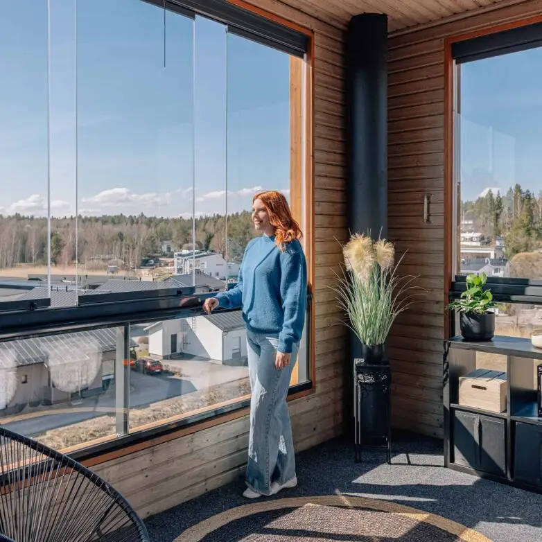 Woman standing on an enclosed balcony with frameless glass walls, enjoying panoramic residential views on a sunny day.