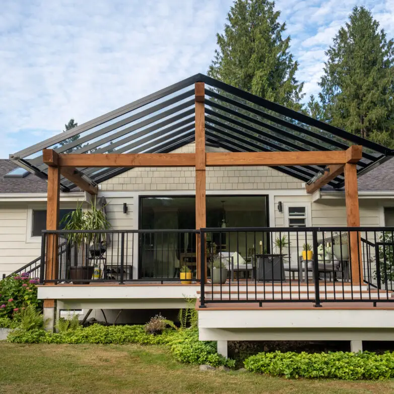 Elevated backyard deck with wood beams and a gabled glass roof structure, overlooking a landscaped garden.
