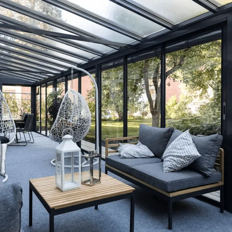 Patio enclosure sunroom with sliding glass walls and a sloped glass roof, featuring modern lounge furniture and garden views.