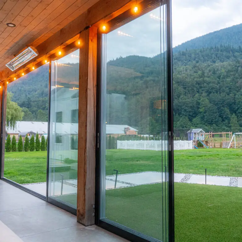 Glass-walled patio enclosure with panoramic lake and forest views, wood decking, and a cozy sofa set.