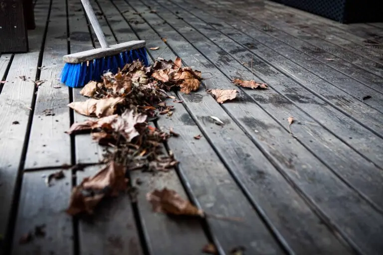 Close-up of a blue broom sweeping dry autumn leaves across a wooden patio deck, showing seasonal outdoor cleaning and maintenance.