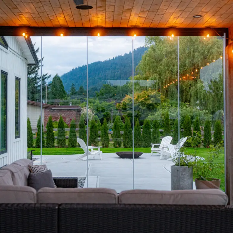 Modern patio enclosure with glass walls offering unobstructed views of the backyard, complete with cozy sectional sofa and string lighting.