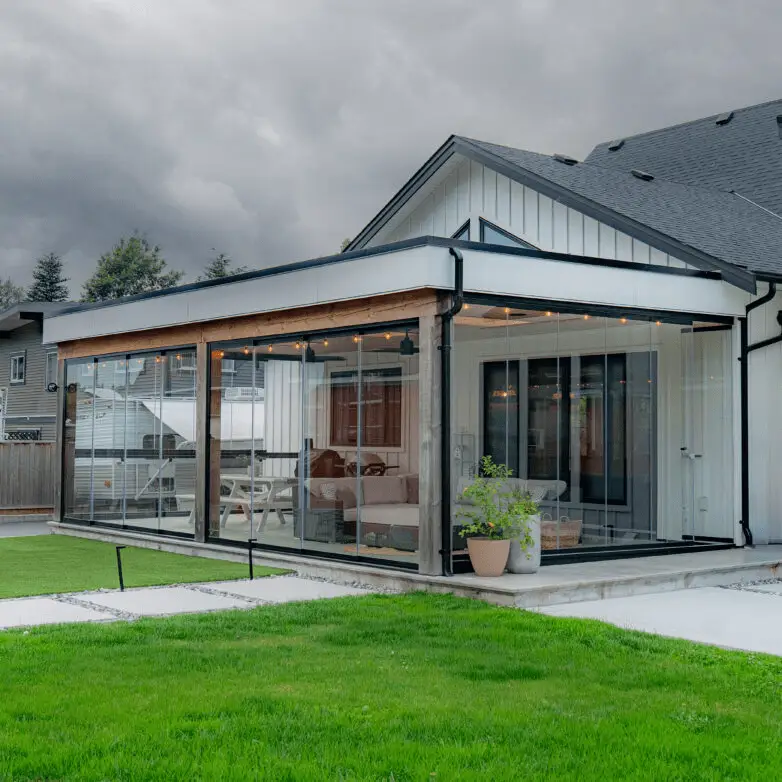 Modern patio enclosure addition with glass walls and flat roof attached to a white house, opening onto a lawn.