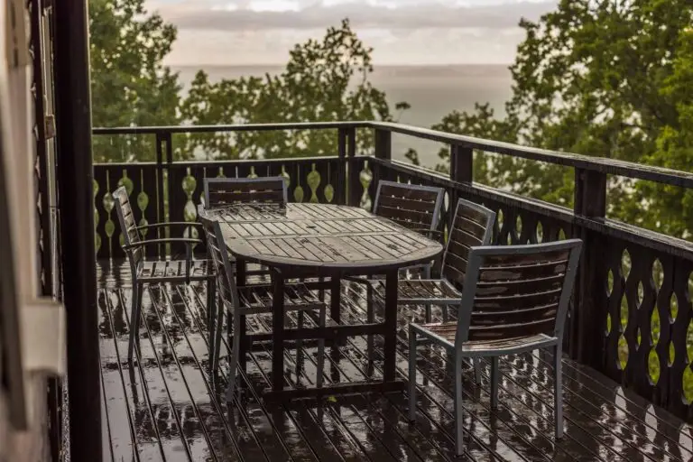 Wooden patio deck with outdoor dining table and chairs glistening after rainfall, overlooking lush trees and a lake view in the distance.