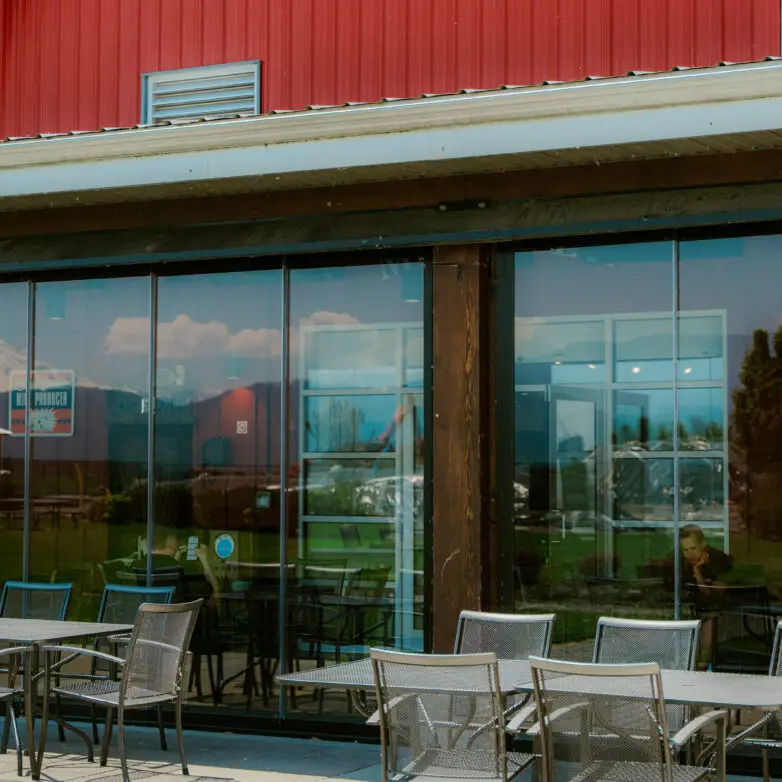 Restaurant exterior with floor-to-ceiling glass walls reflecting blue skies, offering an indoor-outdoor dining area with metal tables and chairs.