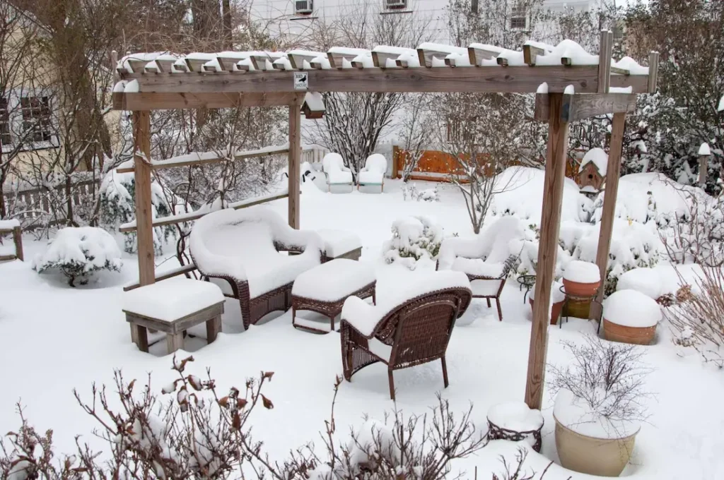Backyard patio furniture and pergola covered in heavy snow, creating a winter garden scene with wicker chairs, potted plants, and frosted trees.