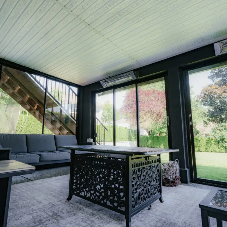 Patio enclosure with black-framed sliding glass doors, gray sofa seating, and an ornate fire table, overlooking a green backyard.