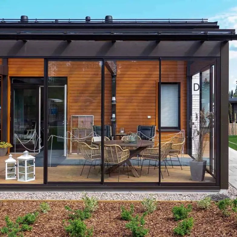 Modern sunroom with black-framed glass walls, sectional seating, and a decorative fire table overlooking a green backyard.