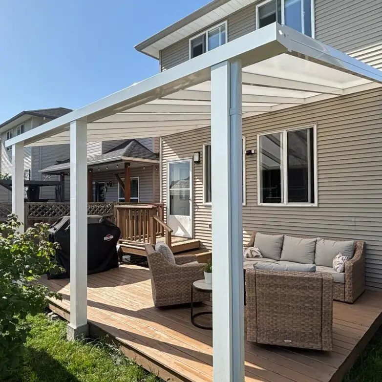 White-framed glass patio cover extending from a suburban home, shading an outdoor seating area with wicker furniture.