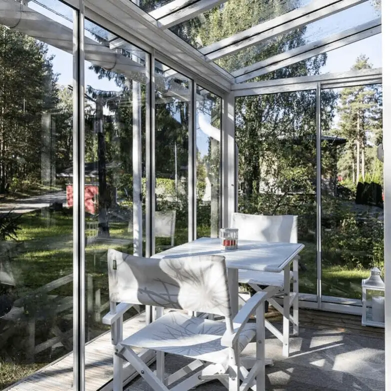 White-framed patio enclosure with glass walls and roof, furnished with a small table and chairs overlooking a wooded yard.